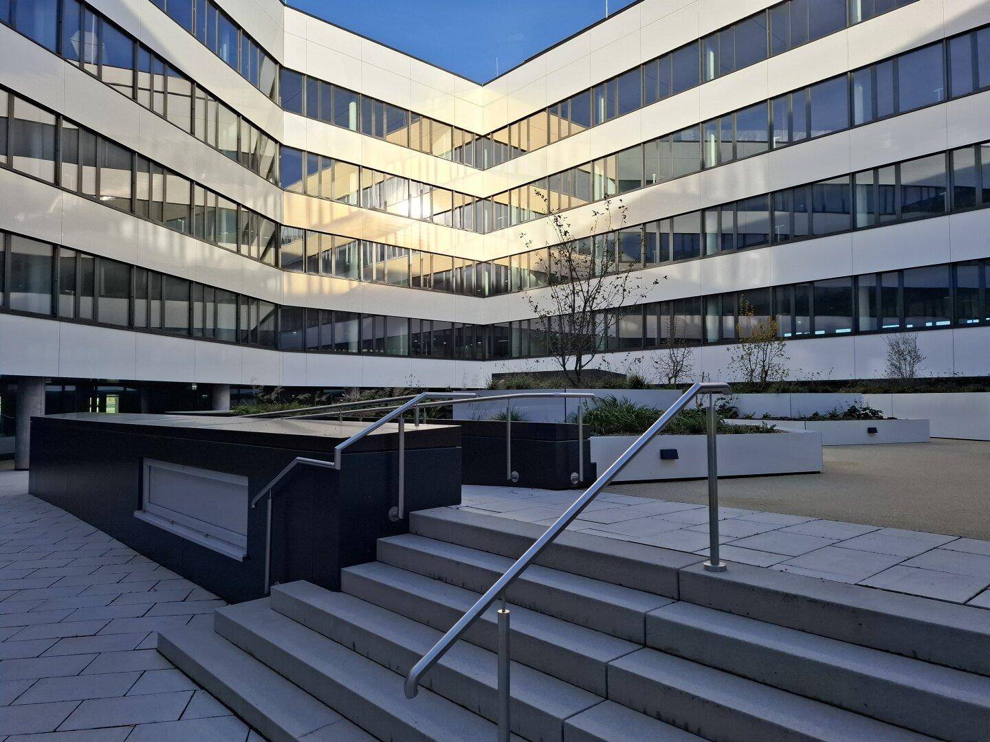 Moderne Bürogebäude-Fassade mit Innenhof, Treppen und begrünten Pflanzbeeten.