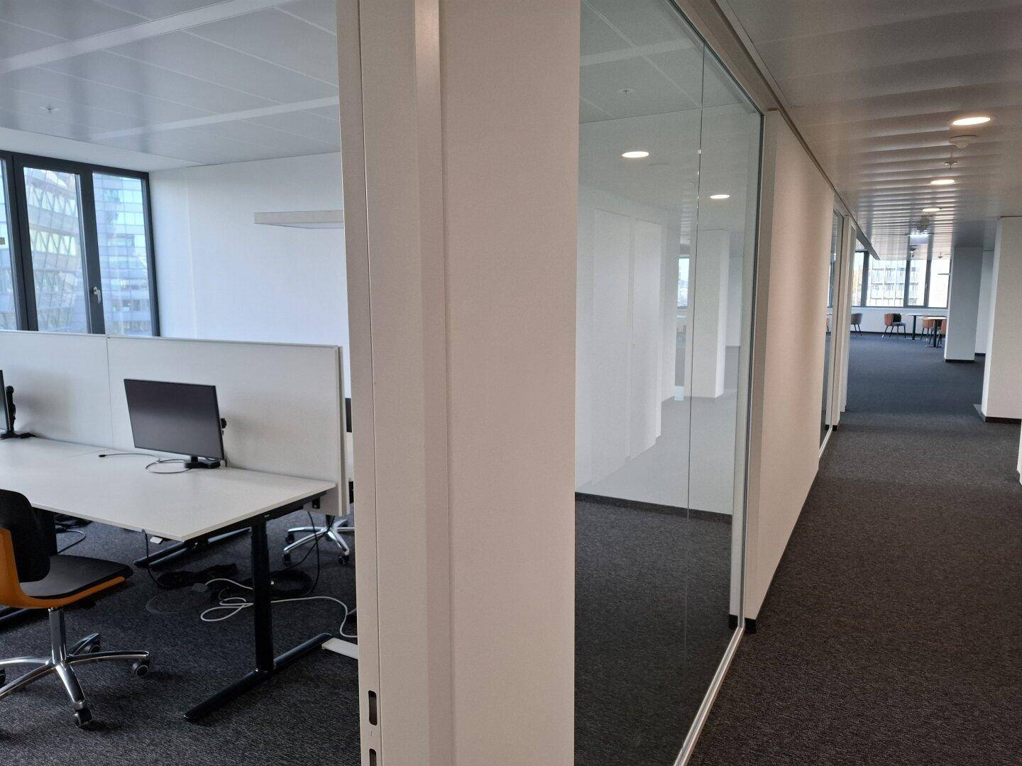Moderner Bürobereich mit Schreibtischen und Monitoren, durch eine Glaswand vom Flur getrennt.