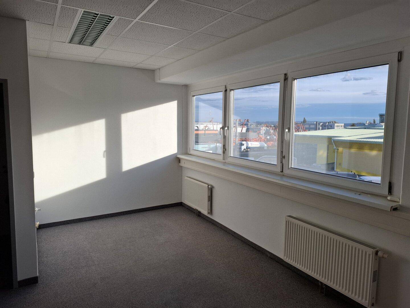 Heller Büroraum mit großen Fenstern, Teppichboden und Heizkörpern, bietet einen weiten Blick.