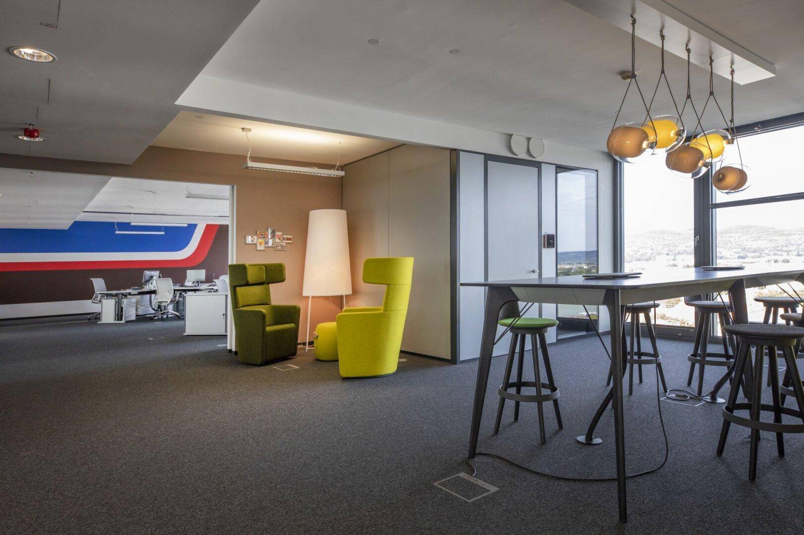 Geräumiger Loungebereich im Büro mit Teppichboden, farbigen Akzenten und großen Fenstern für viel Tageslicht.
