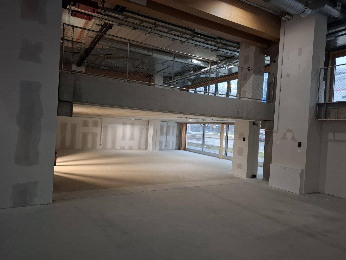 Großer, offener Innenraum im Rohbau mit hohen Decken und großen Fensterfronten zur Straße.