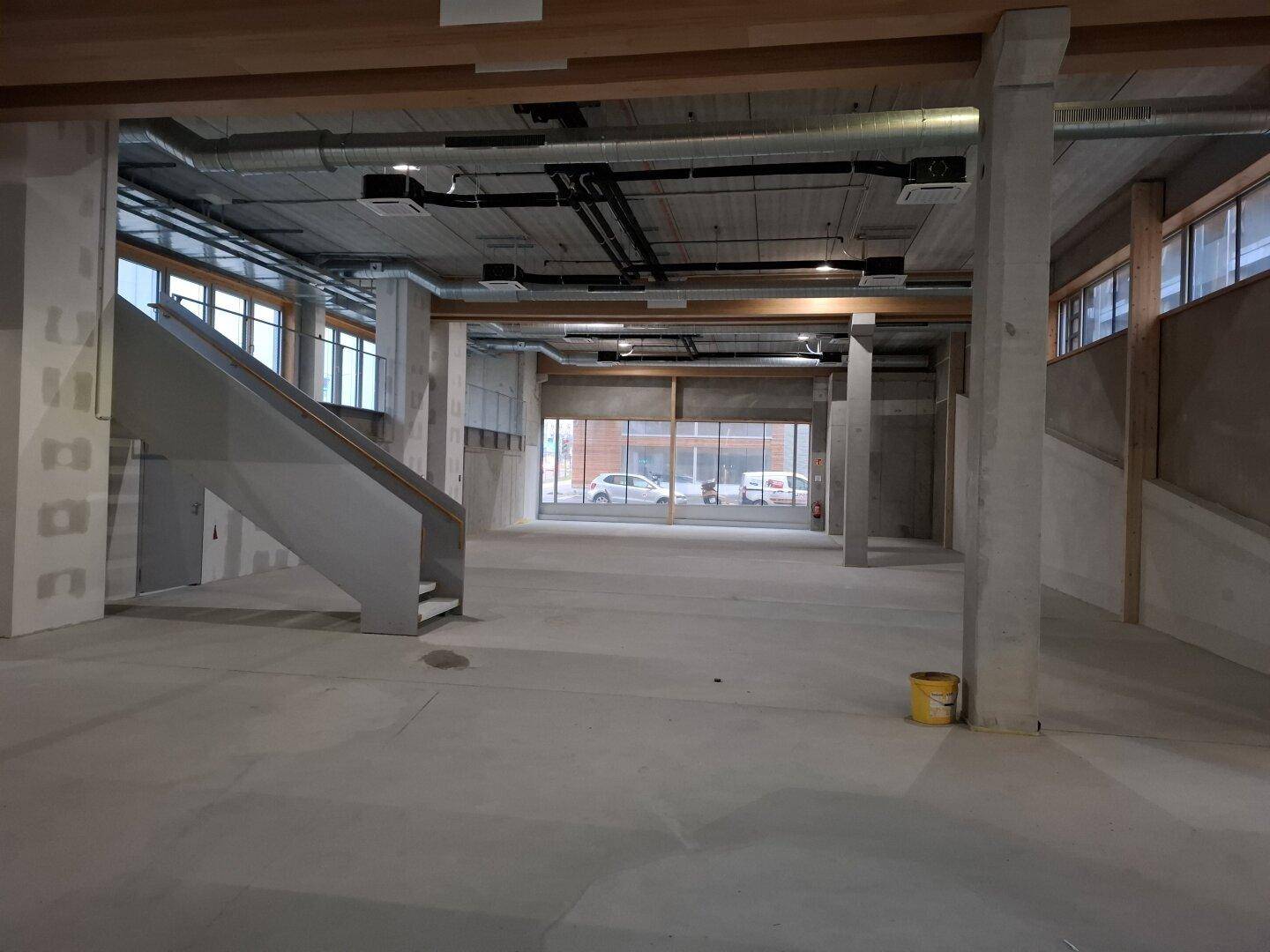 Weitläufiger, unfertiger Gewerberaum mit großen Fenstern, Betonstützen und sichtbaren Installationen.