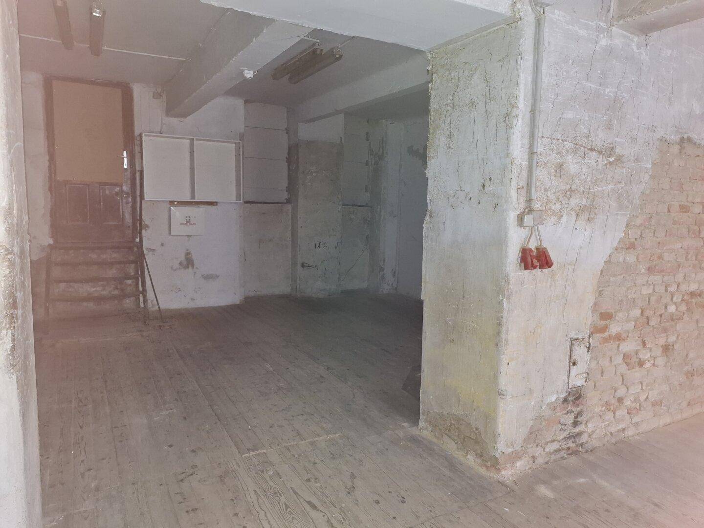 Großer, ungenutzter Raum mit sichtbaren Installationen und einem Zugang zu einem anderen Bereich des Gebäudes.