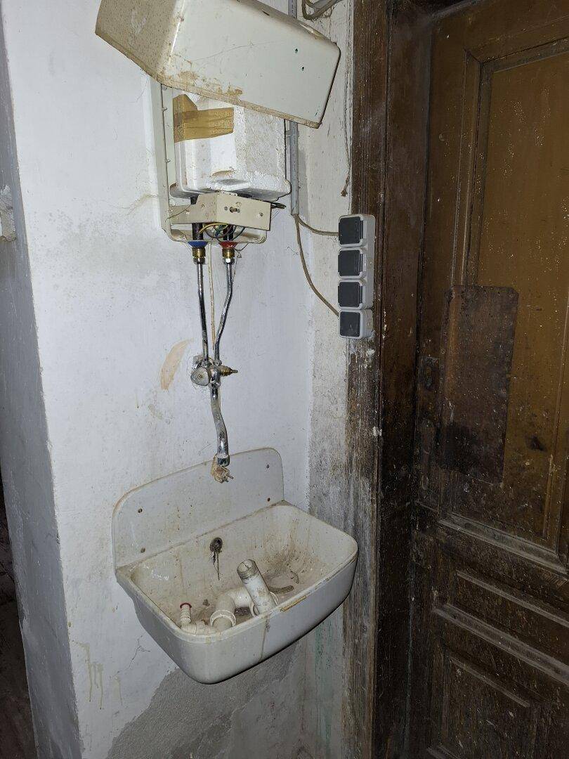 Altes Waschbecken mit Boiler und Steckdosen an einer Wand neben einer abgenutzten Holztür, in schlechtem Zustand.