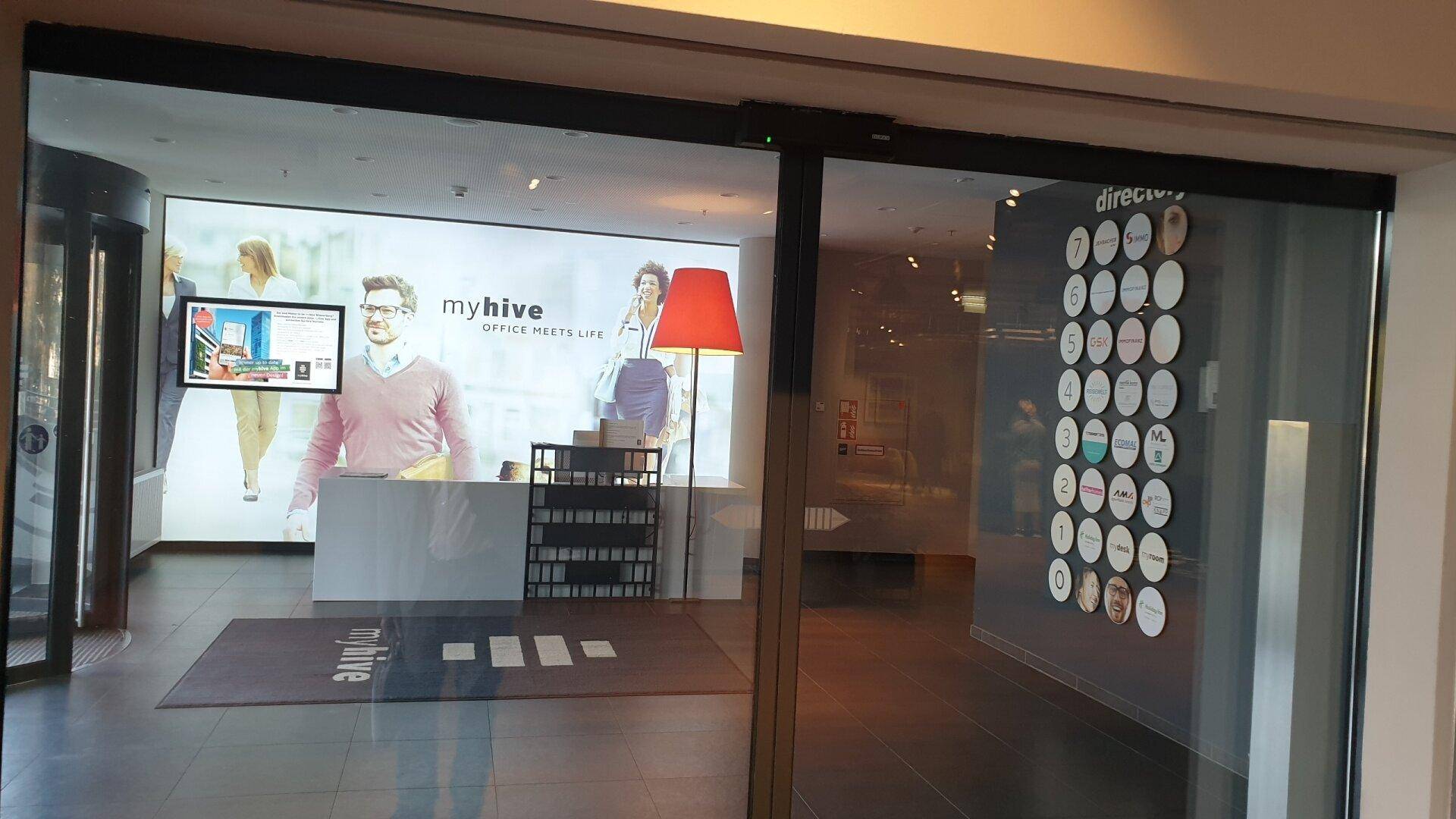 Moderner Eingangsbereich mit Glasfront, digitaler Anzeige und einem großen Bildschirm, der Werbung zeigt.