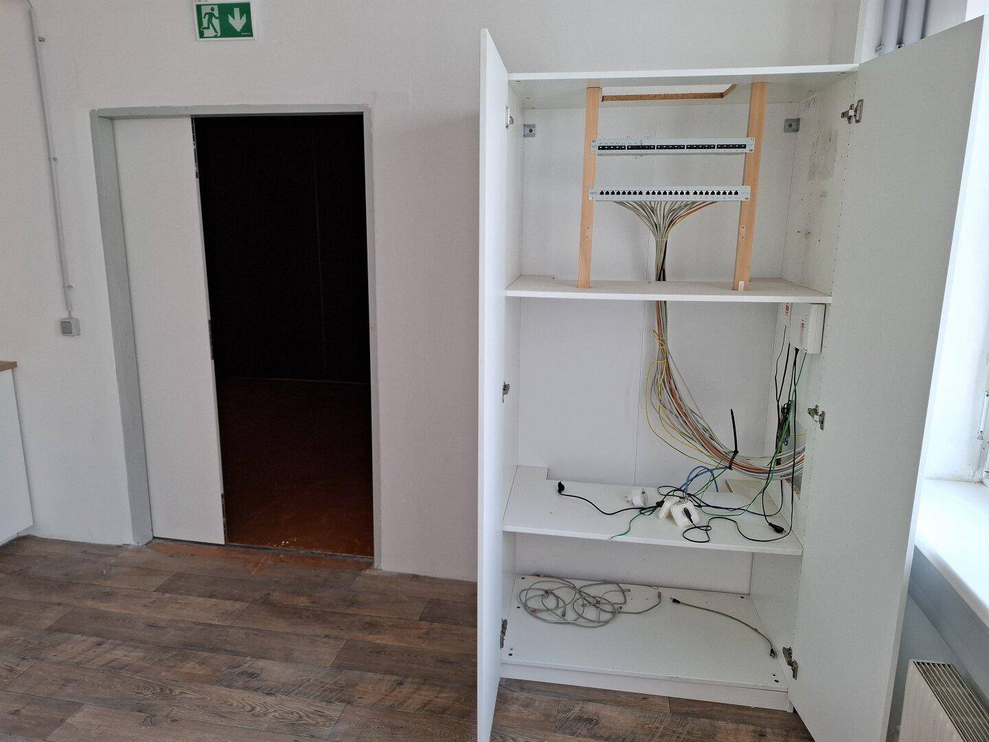 Offener Schrank mit Netzwerkverkabelung und IT-Infrastruktur in einem funktionalen Raum.