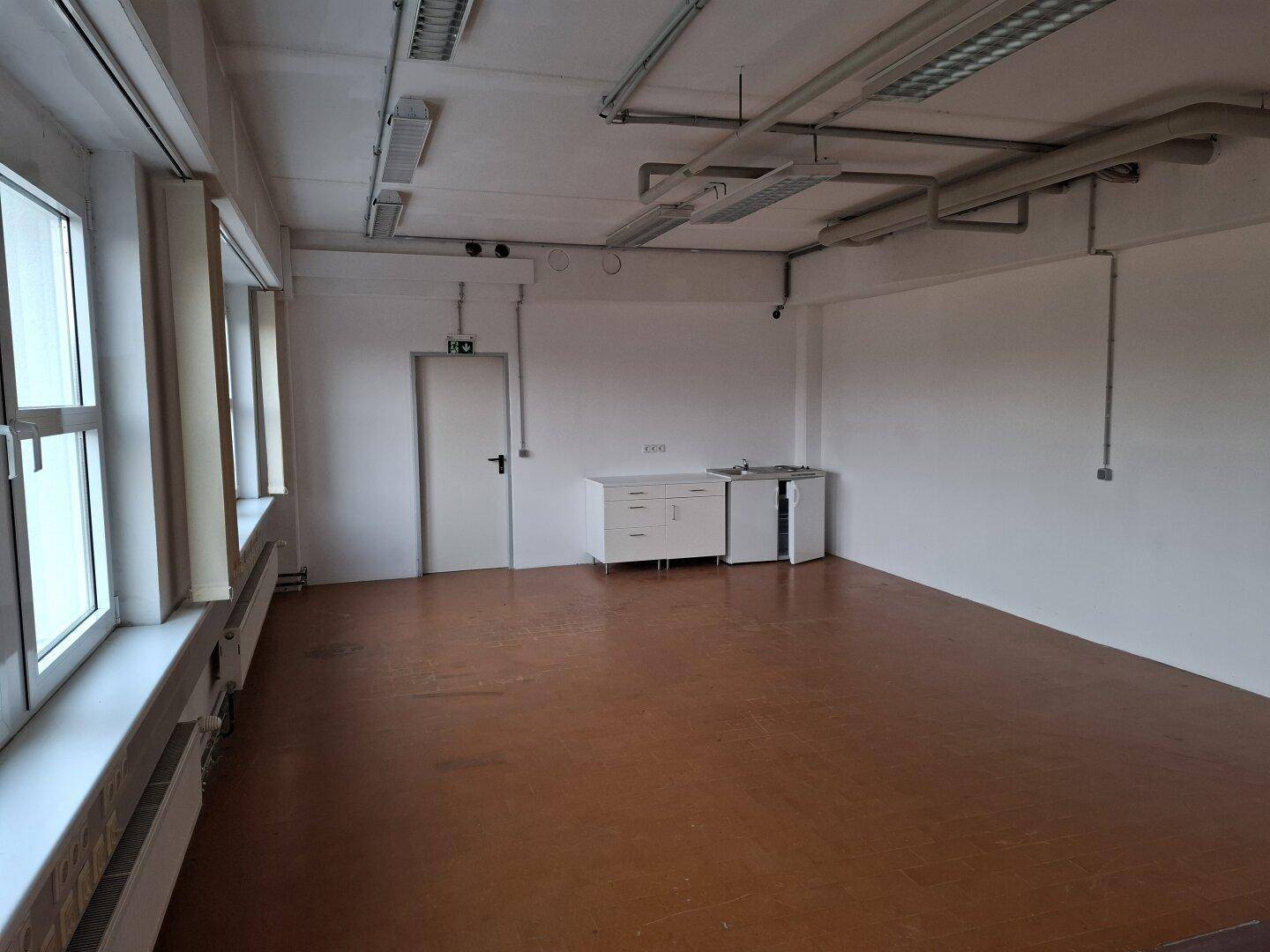 Heller Büroraum mit kleiner Küchenzeile, Fenstern und sichtbaren Deckeninstallationen, ideal für gewerbliche Nutzung.