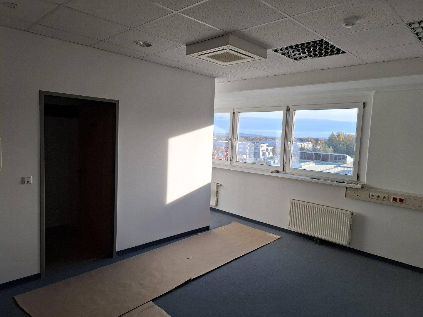 Helles Büro mit großen Fenstern, Teppichboden und Blick auf die Umgebung, bereit zur Einrichtung.