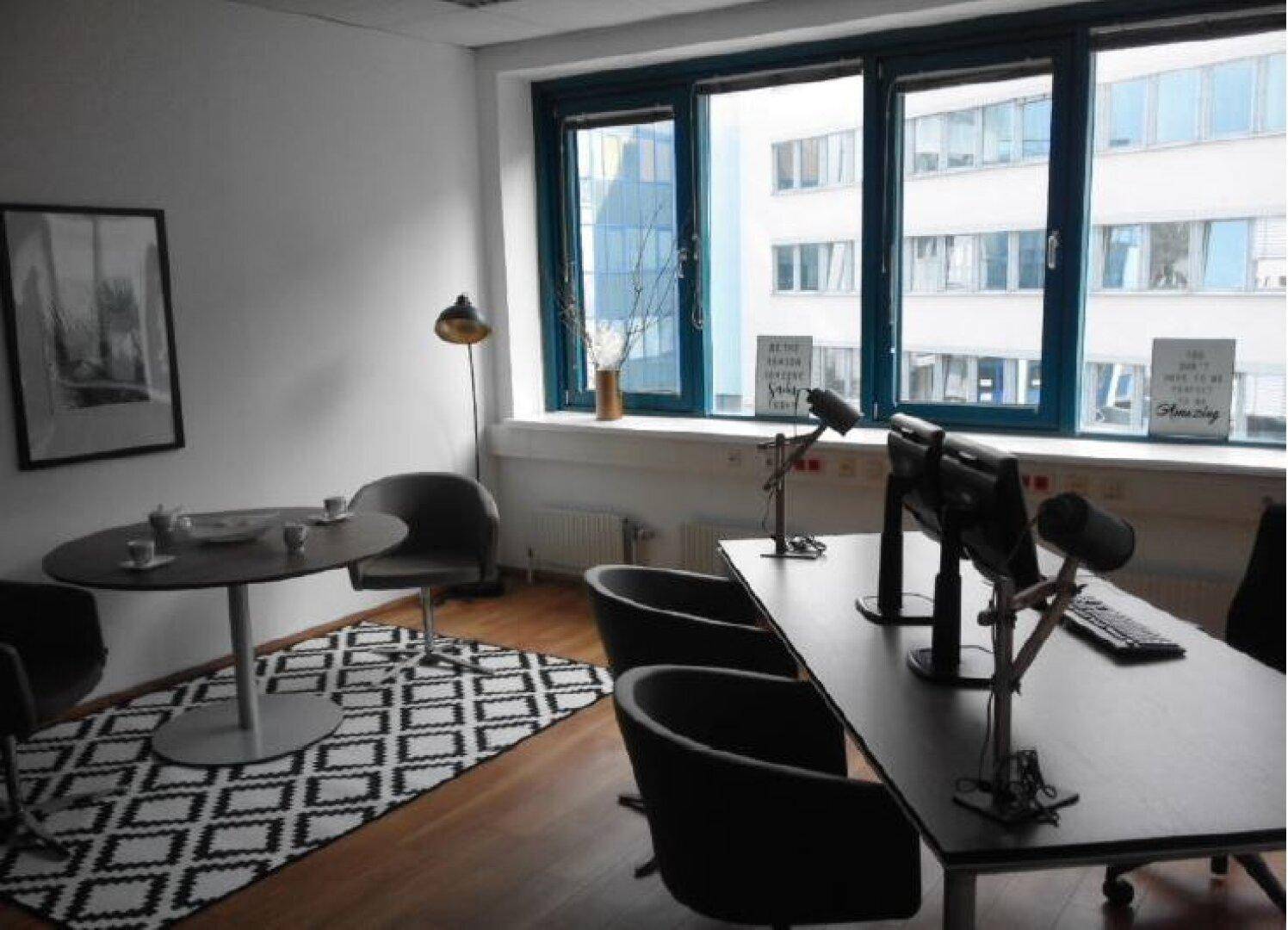 Moderner Büroraum mit Besprechungstisch, Arbeitsplätzen und großen Fenstern, die viel Tageslicht hereinlassen.