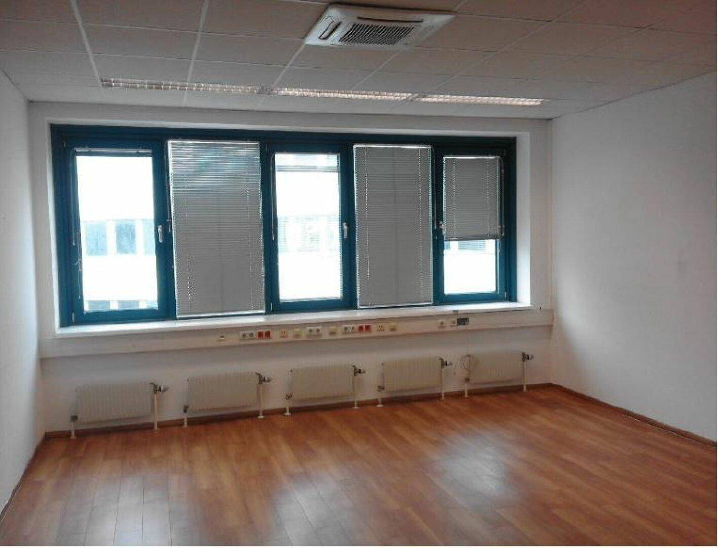 Leerer Büroraum mit großen Fenstern, Jalousien und Heizkörpern unter der Fensterbank, bereit zur Einrichtung.
