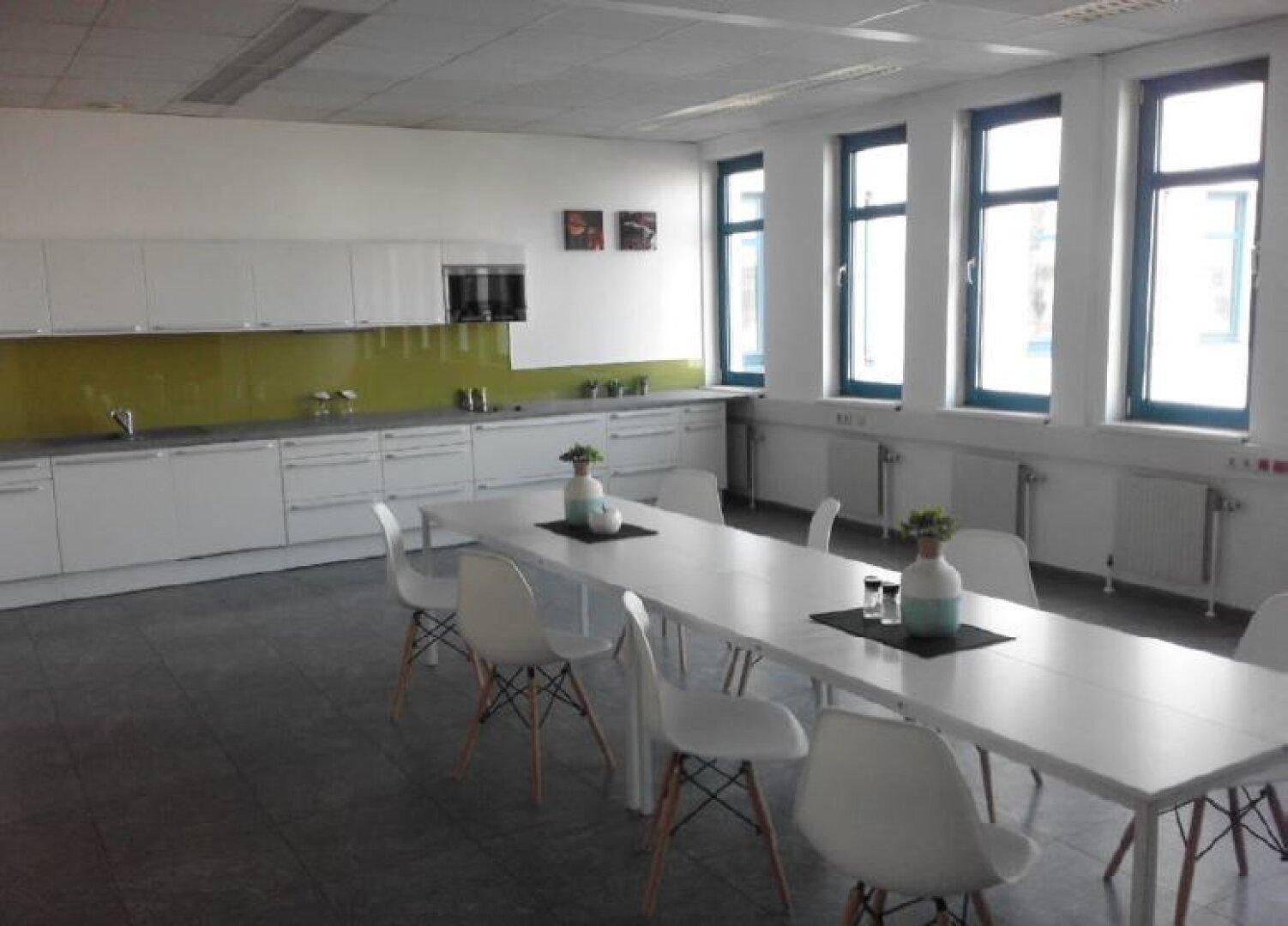 Moderne Gemeinschaftsküche mit langer Tafel, weißen Schränken und grüner Spritzschutzwand, ideal für Pausen.
