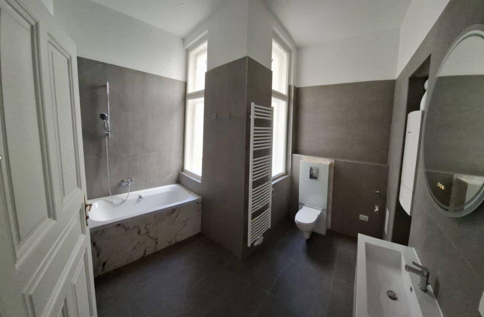 Modernes Badezimmer mit grauen Fliesen, Badewanne, wandhängendem WC und Fenster.