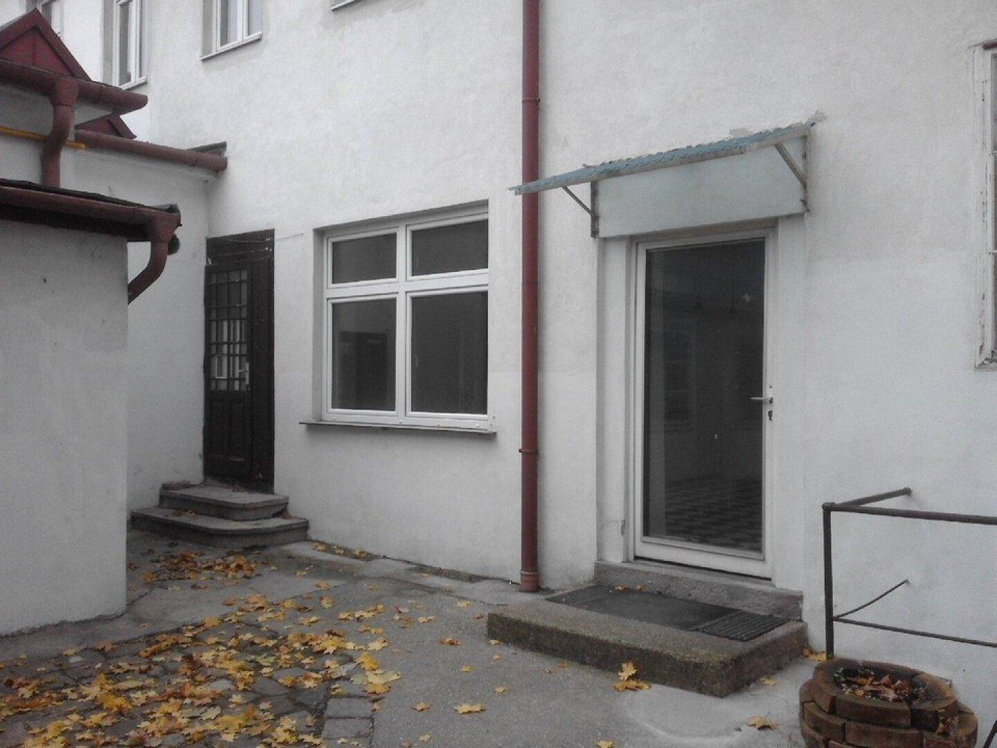 Außenansicht eines Gebäudes mit zwei Eingangstüren, einem Fenster und einem gepflasterten Hofbereich.