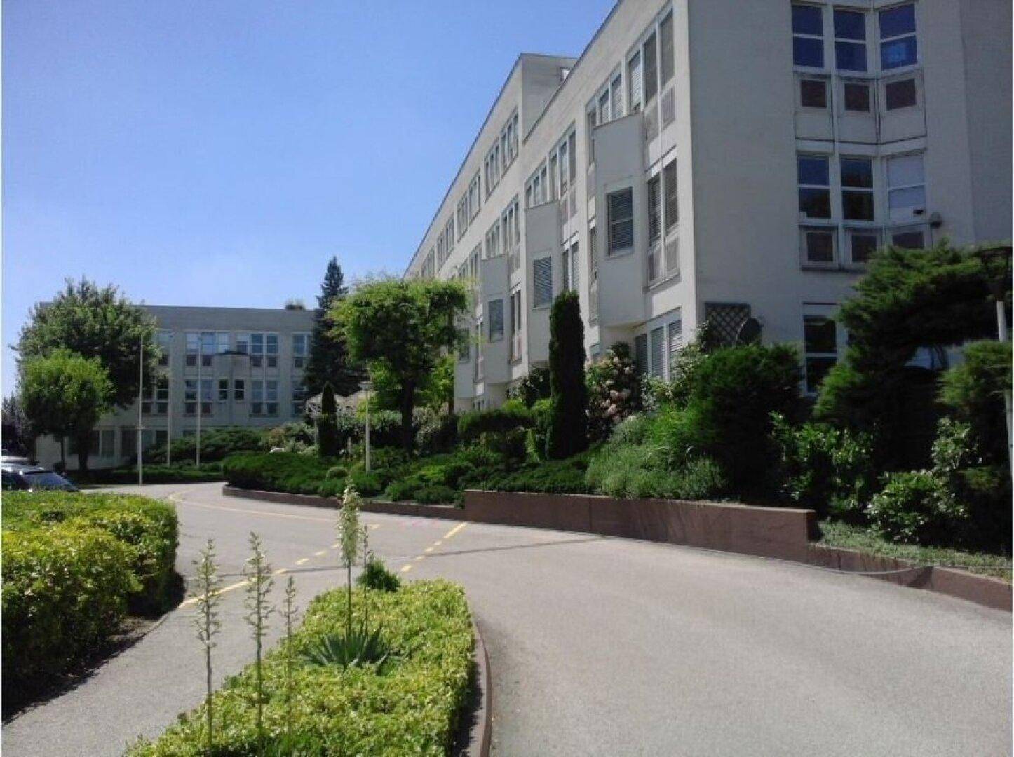 Modernes Gebäude mit vielen Fenstern und gepflegter Grünanlage an einem sonnigen Tag, umgeben von Bäumen.