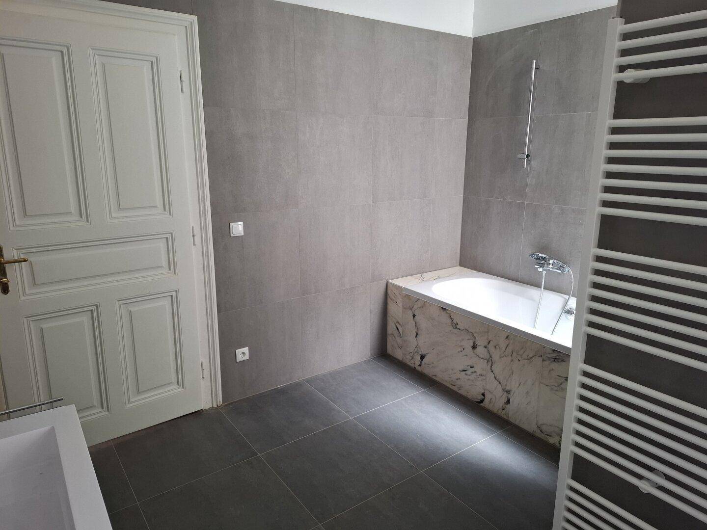 Modernes Badezimmer mit grauen Fliesen, einer Badewanne mit Marmorverkleidung und Handtuchheizkörper.