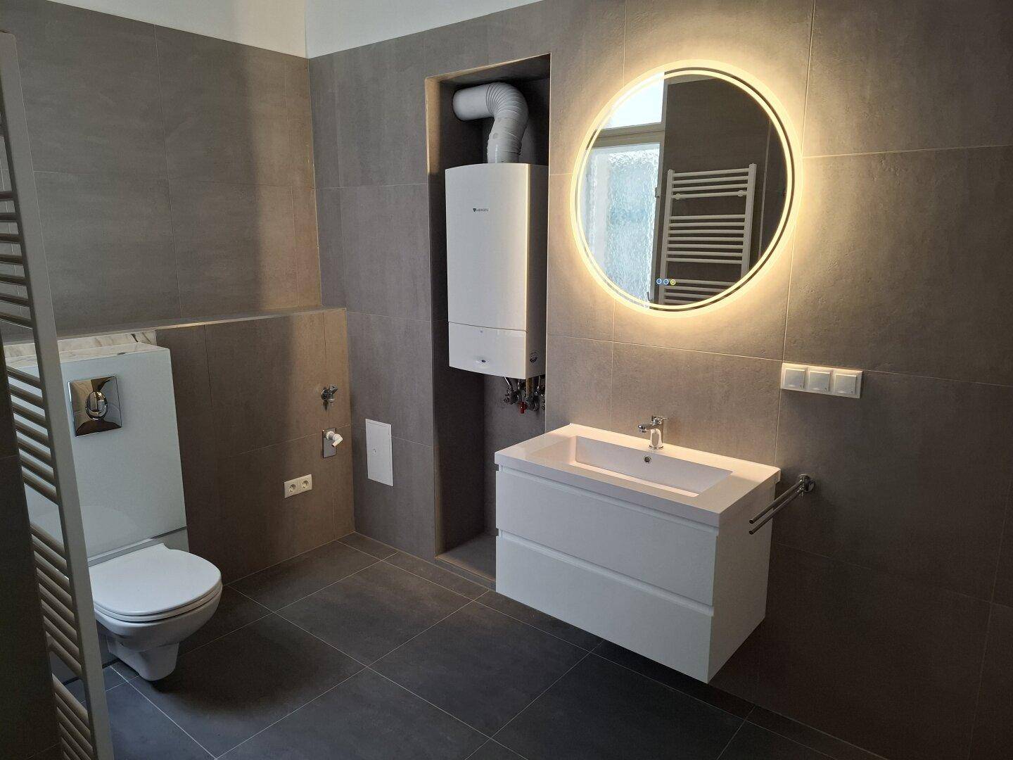 Zeitgemäßes Badezimmer mit Wand-WC, modernem Waschtisch, beleuchtetem Spiegel und integriertem Warmwasserbereiter.