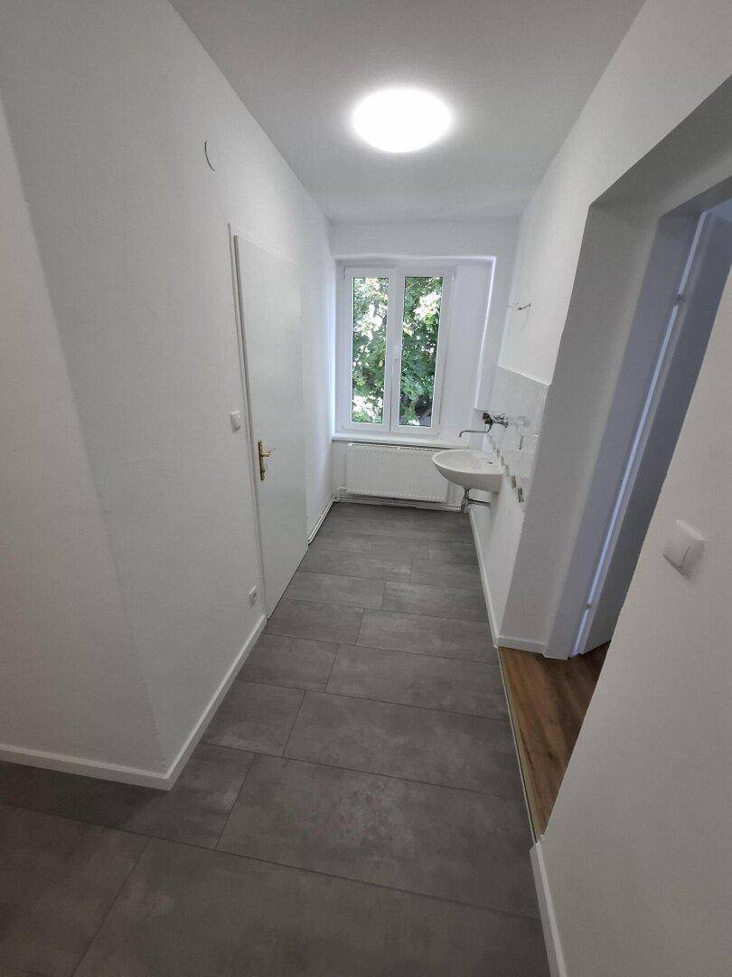 Langer Flur mit grauen Fliesen, weißen Wänden, einem Fenster und einem Waschbecken an der Wand.