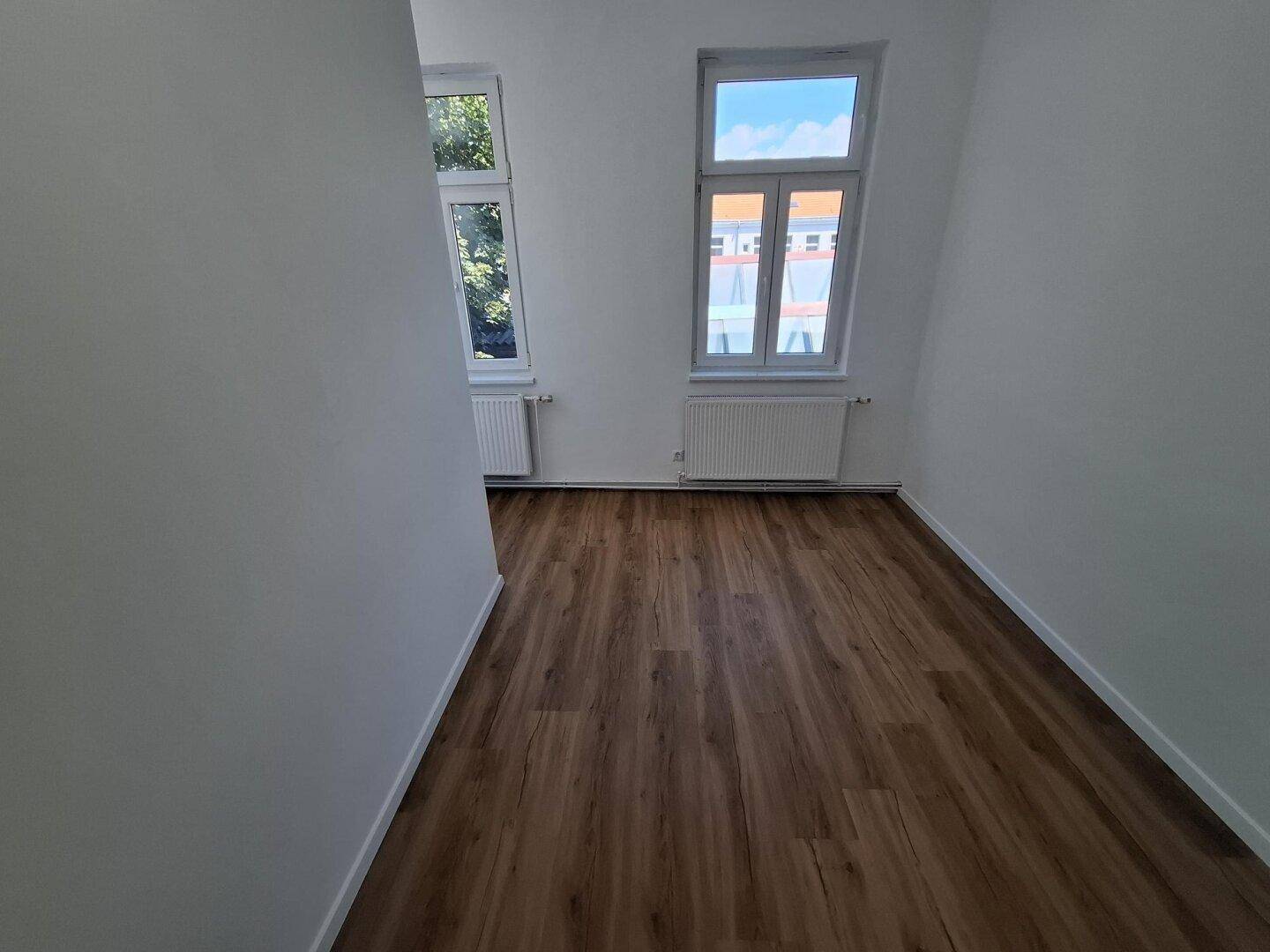 Leerer Raum mit Laminatboden, weißen Wänden und zwei Fenstern, die viel Tageslicht hereinlassen.