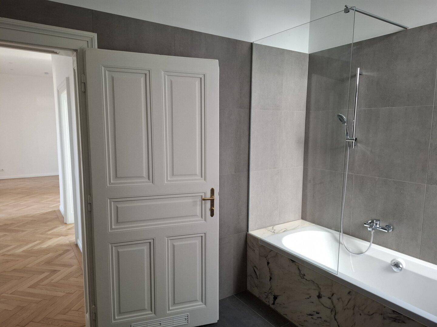 Modernes Badezimmer mit grauen Fliesen, einer Badewanne und einer Duschabtrennung aus Glas.