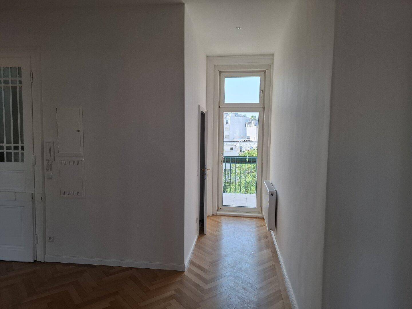 Langer Flur mit Fischgrätparkettboden, der zu einer Tür mit französischem Balkon führt und natürliches Licht spendet.