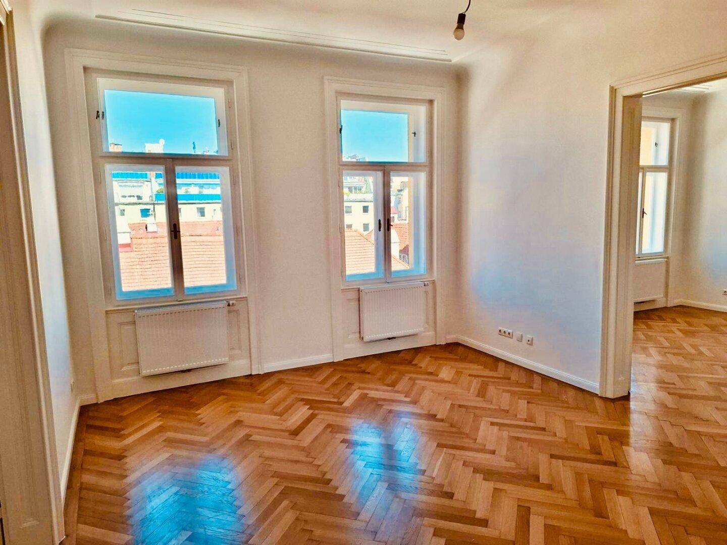 Helles Zimmer mit hohen Fenstern, Fischgrätparkett und Blick auf die umliegenden Dächer.