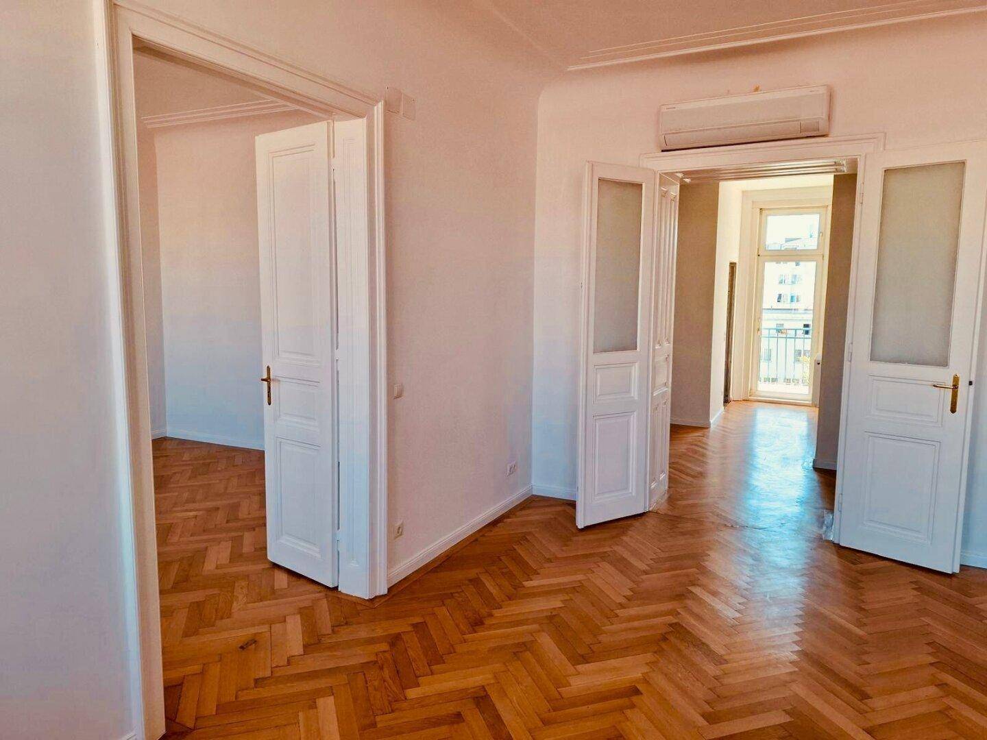 Heller Wohnraum mit Fischgrätparkett, Klimaanlage und Zugang zu einem französischen Balkon.