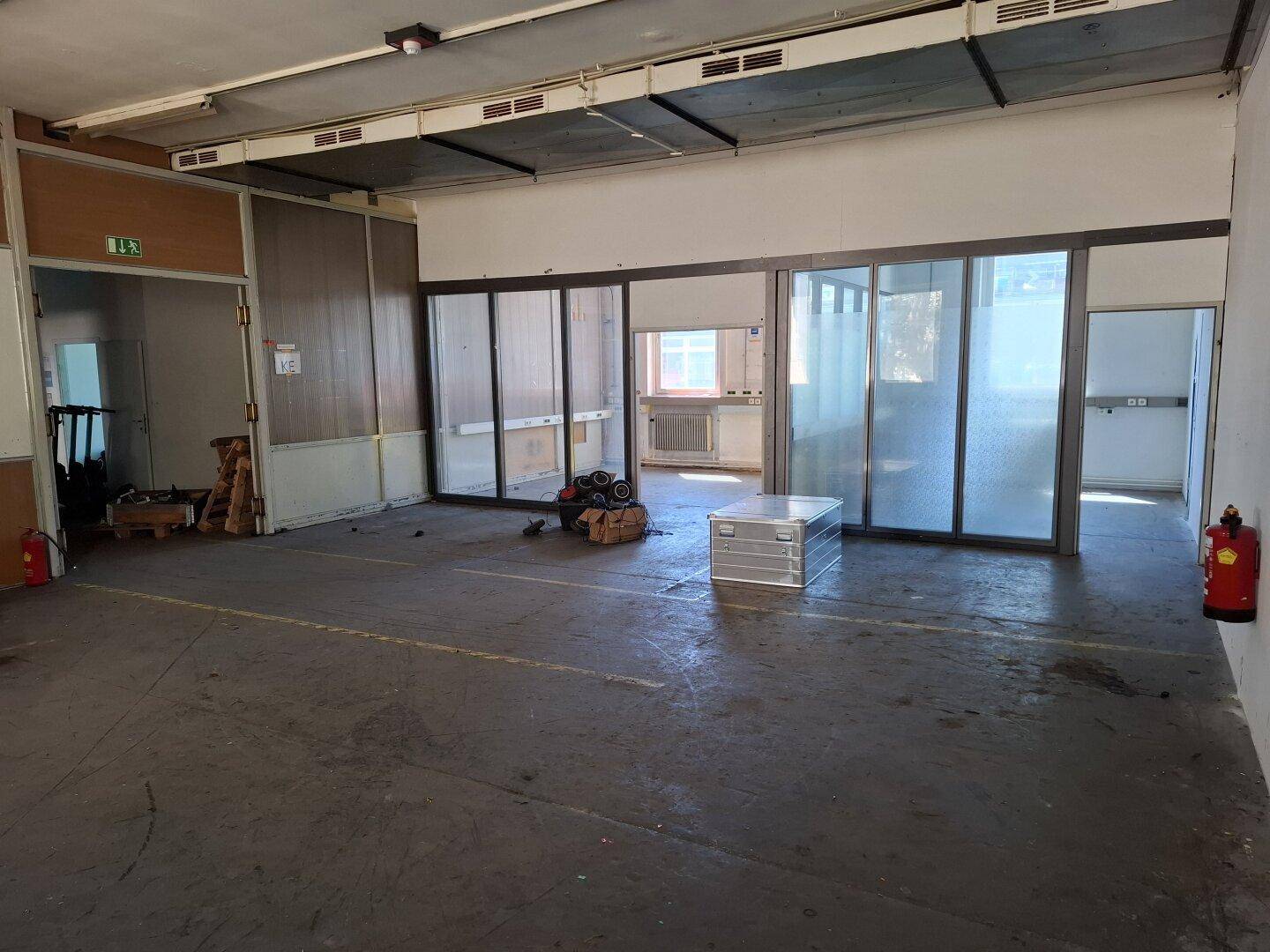 Weiterer Blick in den großen Industrieraum mit Betonboden, Trennwänden und Feuerlöschern an der Wand.