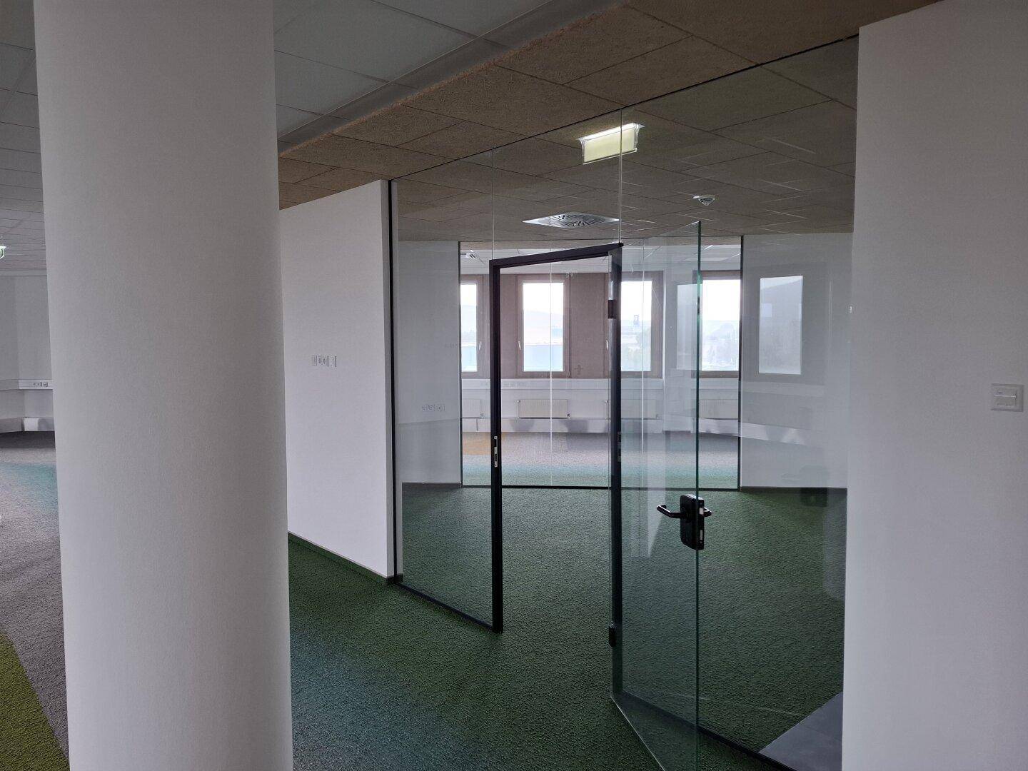 Moderner Bürobereich mit Glaswänden und Teppichboden, der eine helle und offene Arbeitsumgebung schafft.