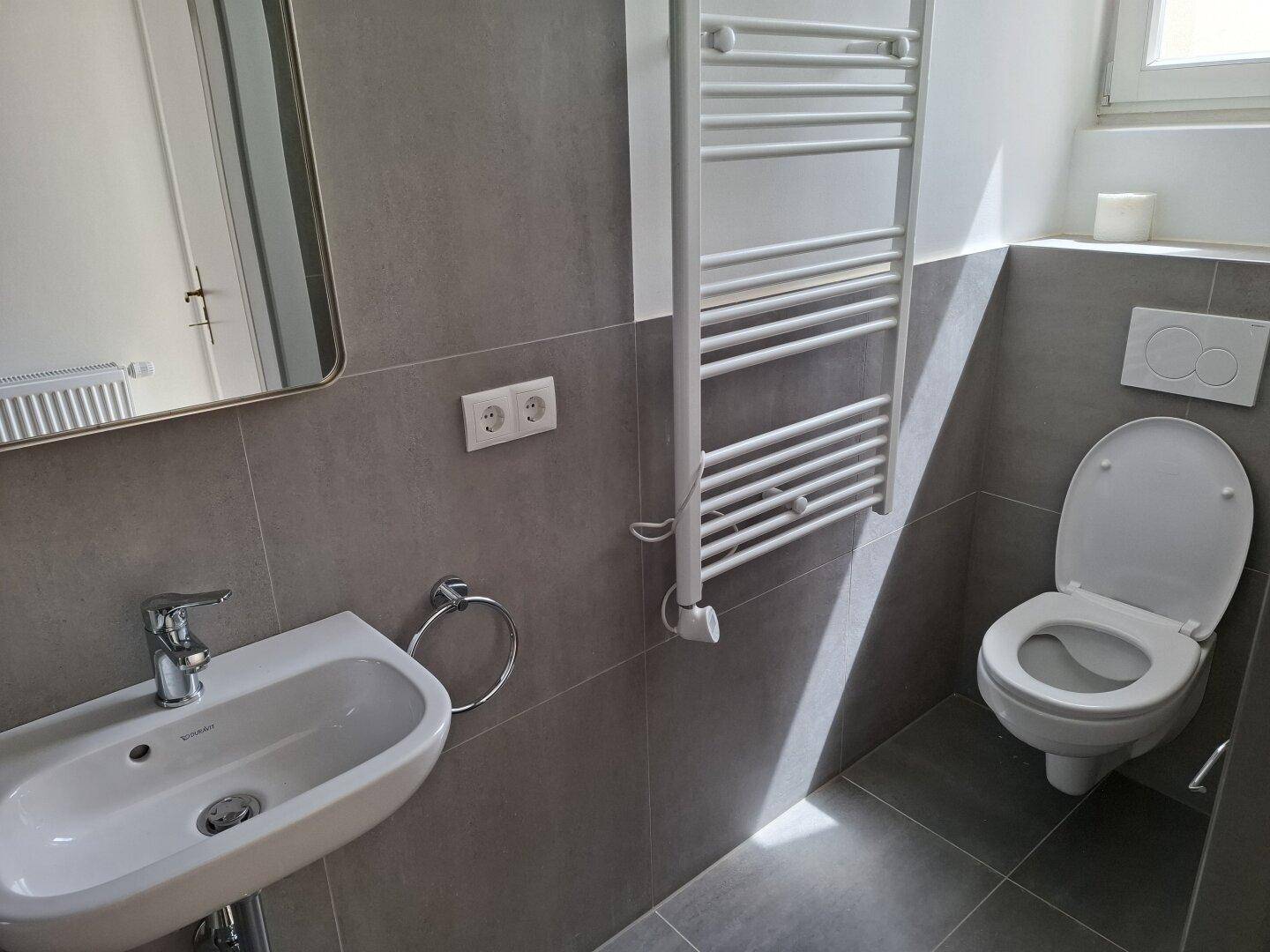 Helles Badezimmer mit modernem WC, Waschbecken, Spiegel und beheiztem Handtuchhalter.