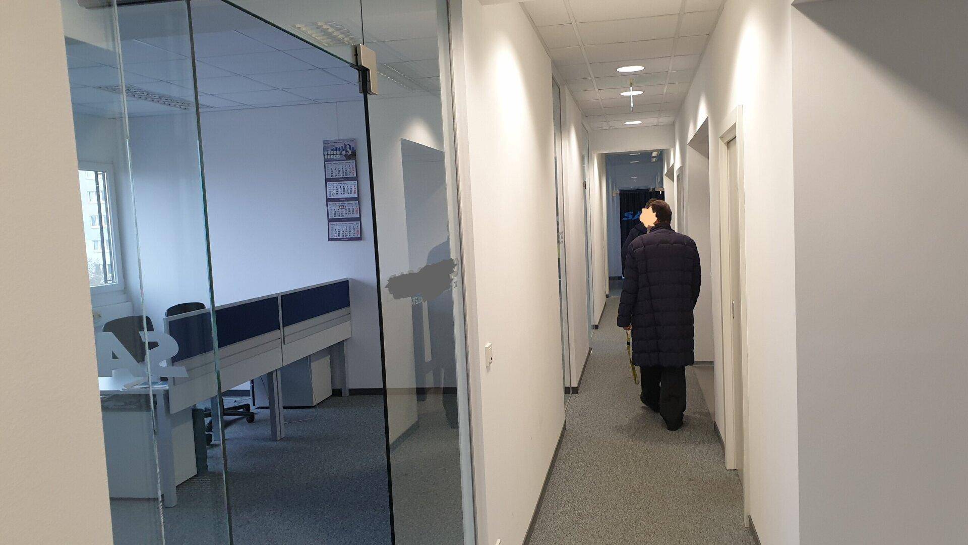Heller Büroflur mit Teppichboden und Glaswänden, die zu den angrenzenden Büroräumen führen.