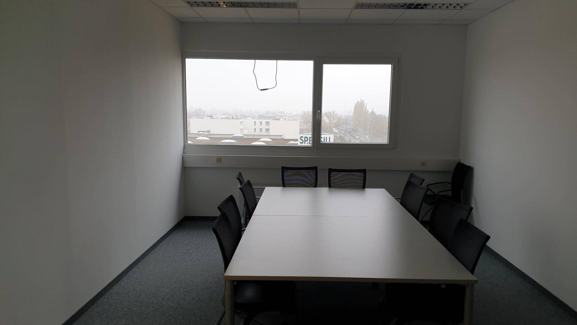Konferenzraum mit großem Tisch und Stühlen, hellen Wänden und Fenstern mit Blick auf die Stadt.