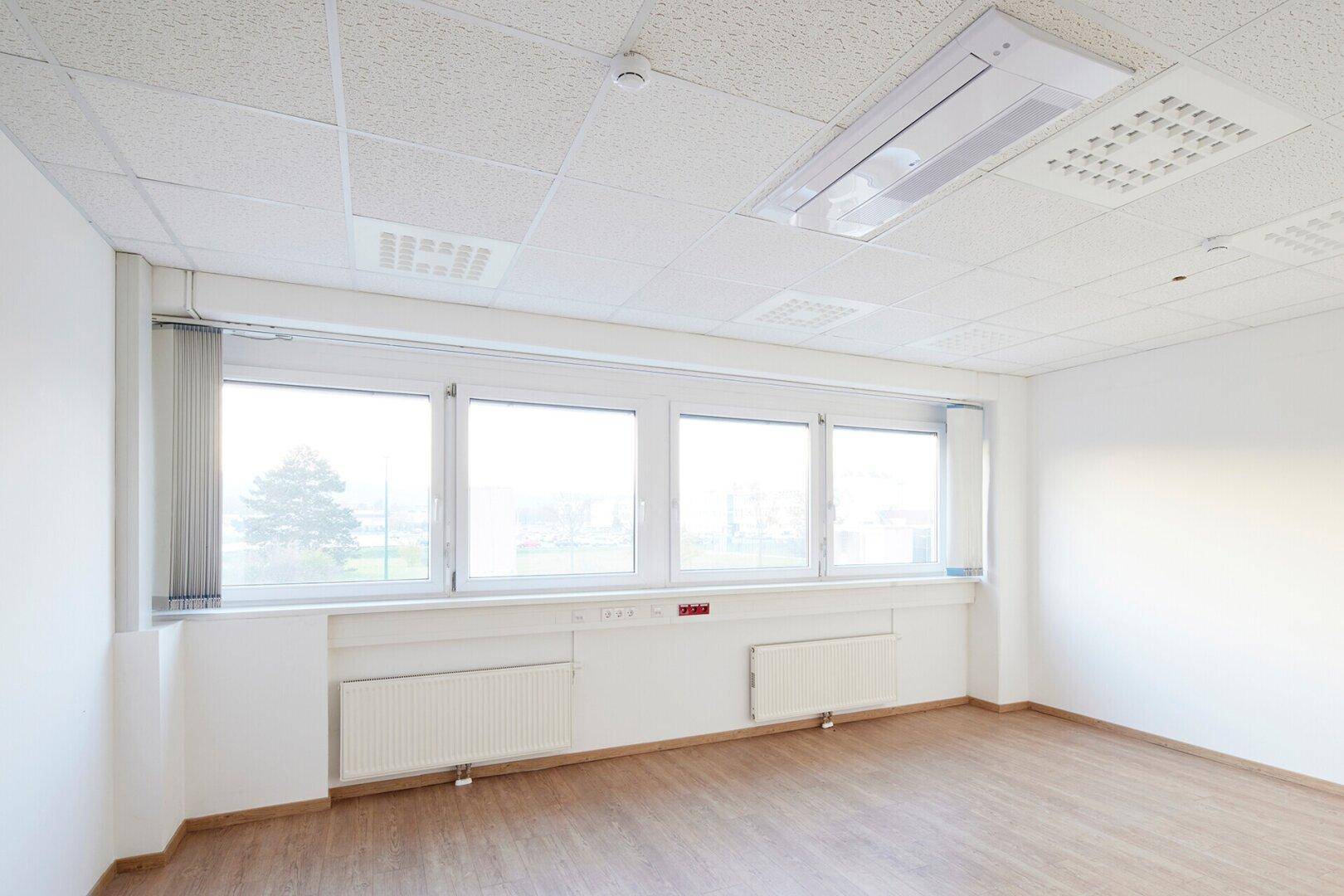 Helles, leeres Büro mit großen Fenstern, Heizkörpern und Holzoptik-Laminatboden.