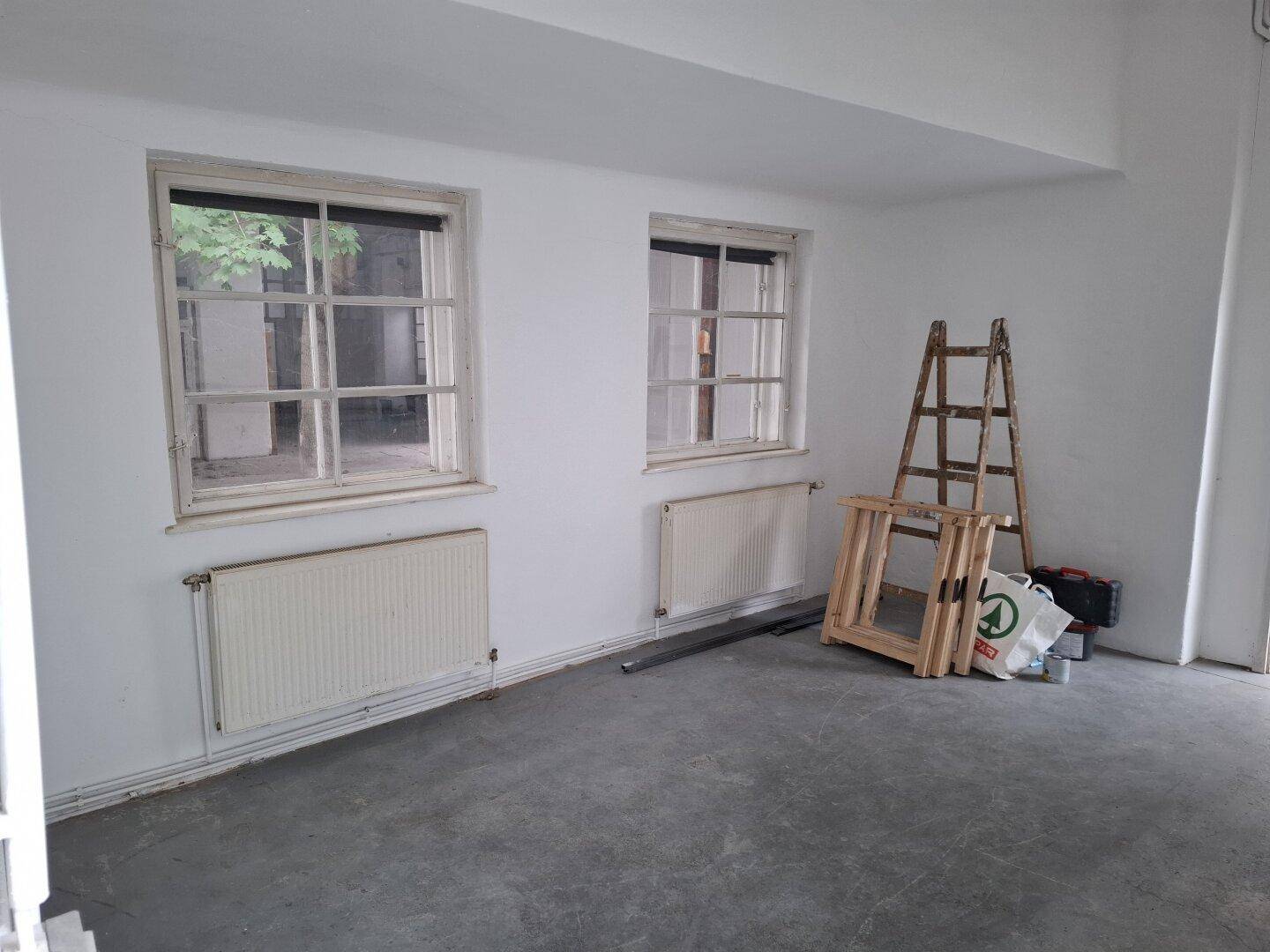 Heller Raum mit zwei Fenstern, Heizkörpern und Betonboden, derzeit als Lagerfläche genutzt.