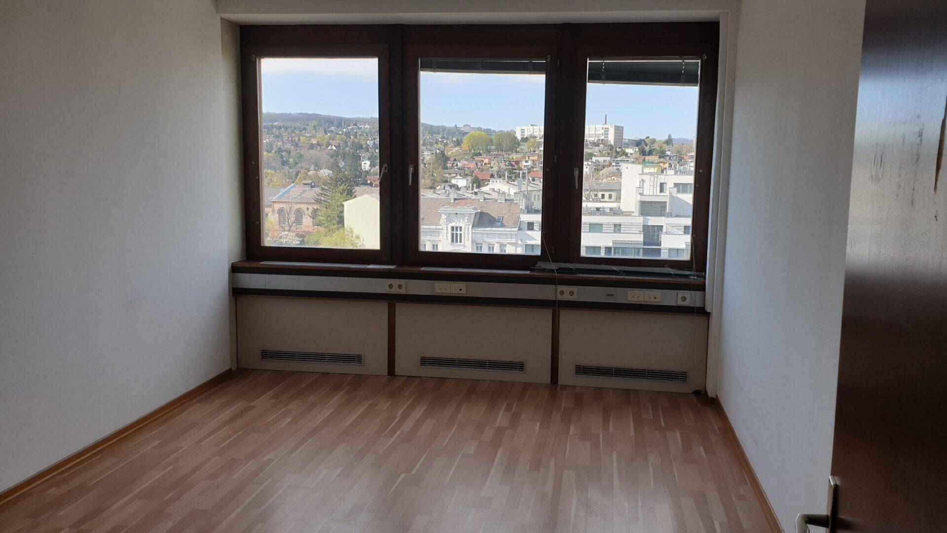 Leerer Büroraum mit Holzboden und breiten Fenstern, die einen weiten Blick über die Stadt bieten.
