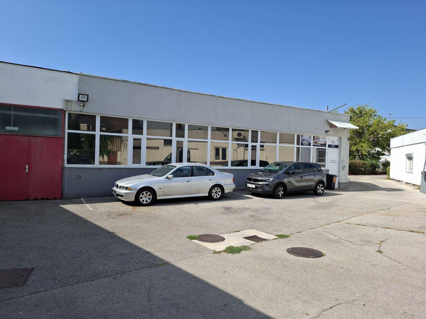 Frontansicht des Gebäudes mit großen Fensterfronten und zwei geparkten Autos auf dem Vorplatz.