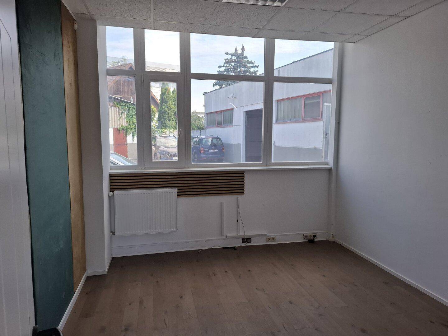 Helles Büro mit Holzboden, großen Fenstern und einem Heizkörper unter einer Holzverkleidung.