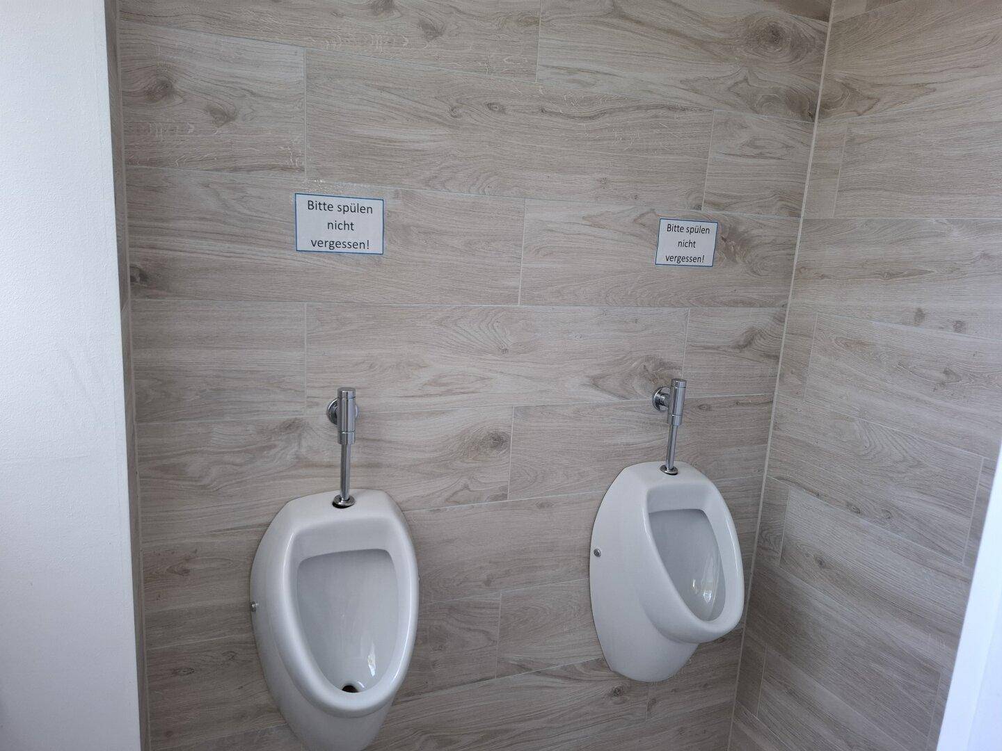 Moderner Herrenwaschraum mit zwei Urinalen und holzgemusterten Fliesen an den Wänden.