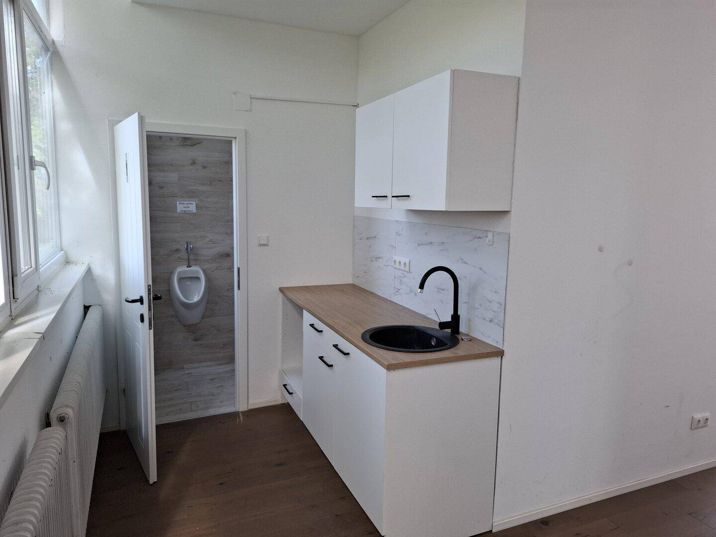 Kleine Küchenzeile mit weißen Schränken, Holztheke und Blick in einen Waschraum mit Urinal.
