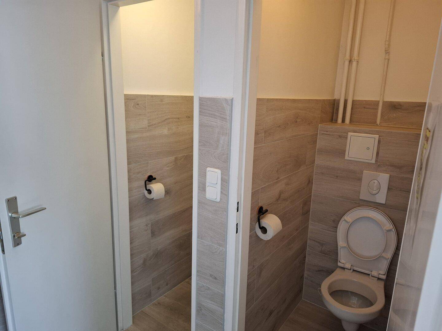 Zwei separate Toilettenkabinen mit holzgemusterten Fliesen und weißen Toiletten.
