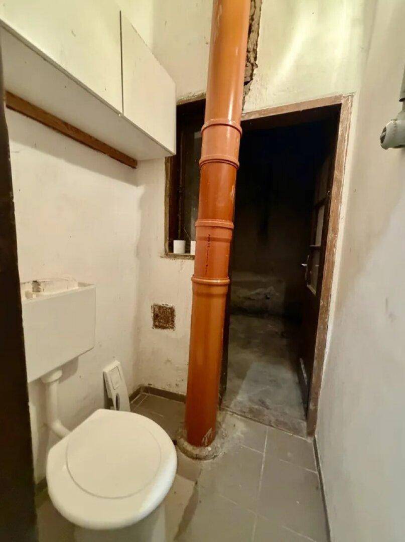 Kleiner, einfacher Raum mit Toilette und sichtbaren Rohren, der eine Renovierung benötigt.