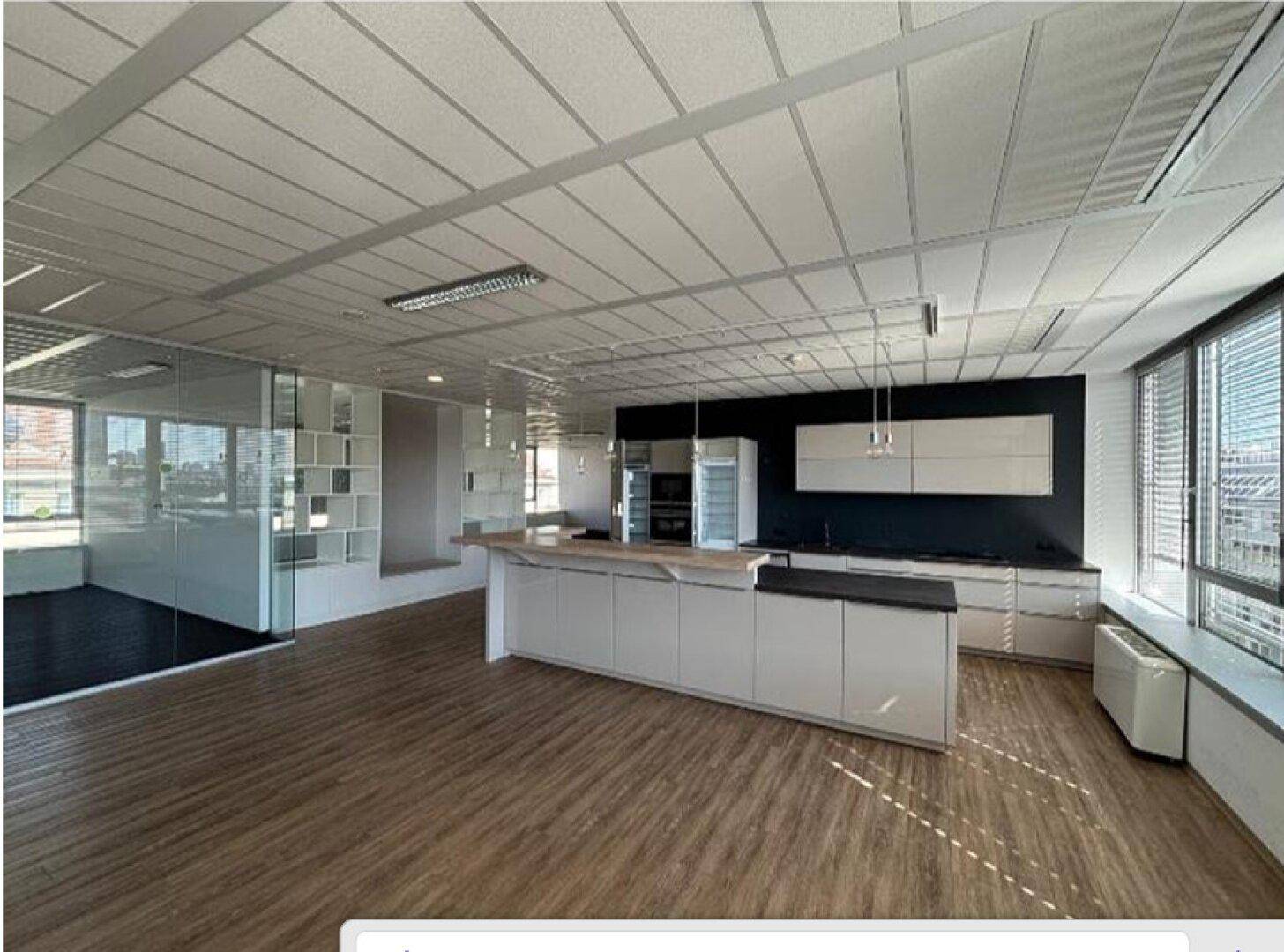 Großzügiger Büroraum mit integrierter moderner Küche, Holzboden und großen Fenstern mit Stadtblick.