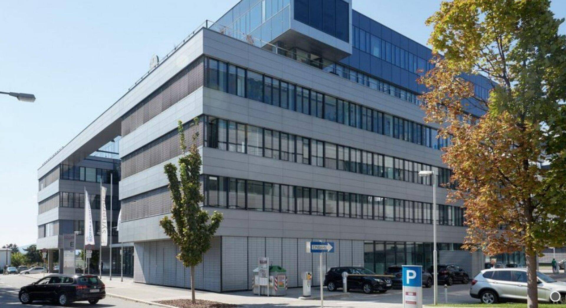 Modernes Bürogebäude mit Glasfassade, mehreren Etagen und Parkplätzen im Freien unter blauem Himmel.