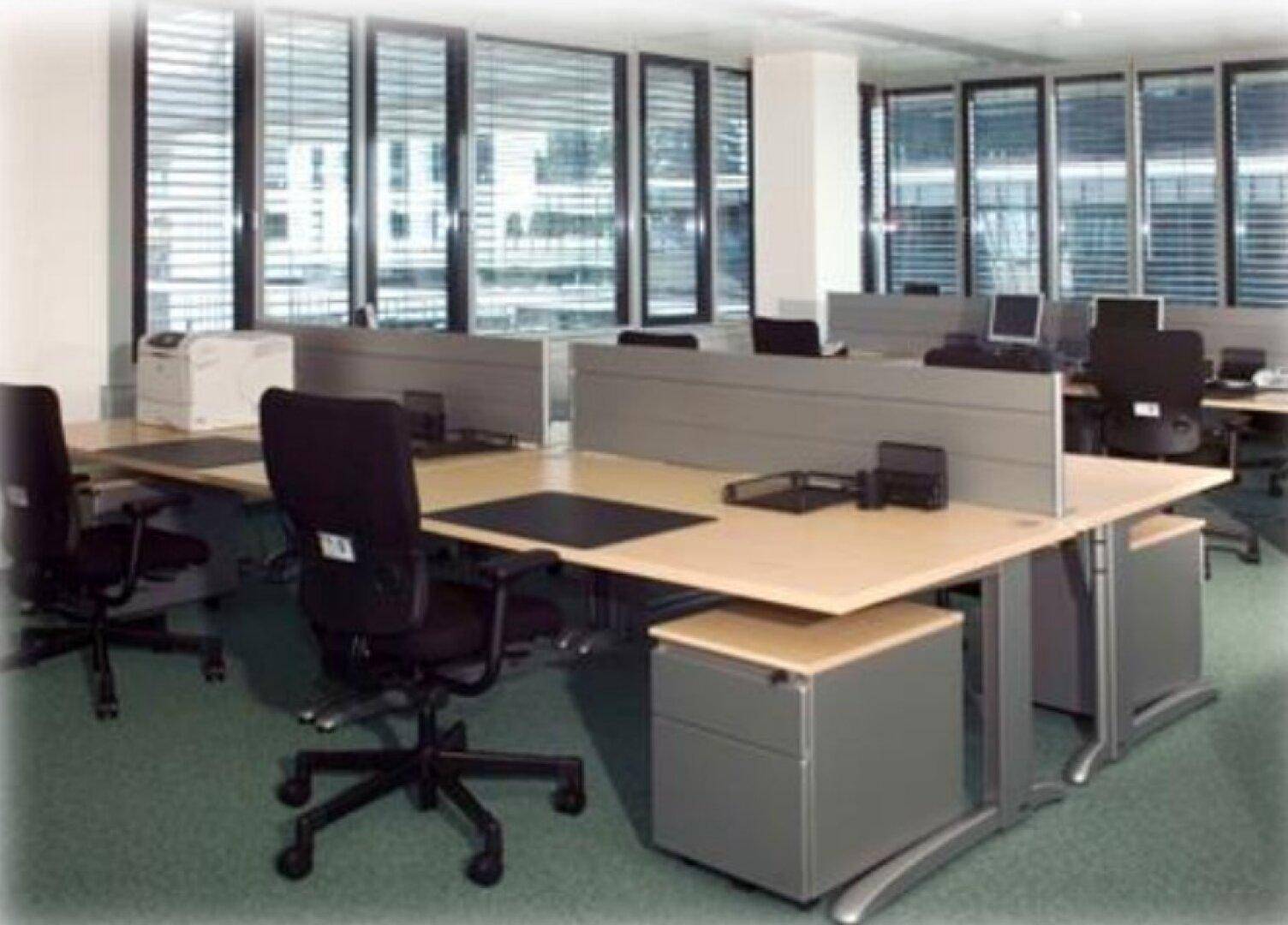 Möblierter Büroraum mit mehreren Arbeitsplätzen, Schreibtischen und Stühlen, ausgestattet mit Jalousien.