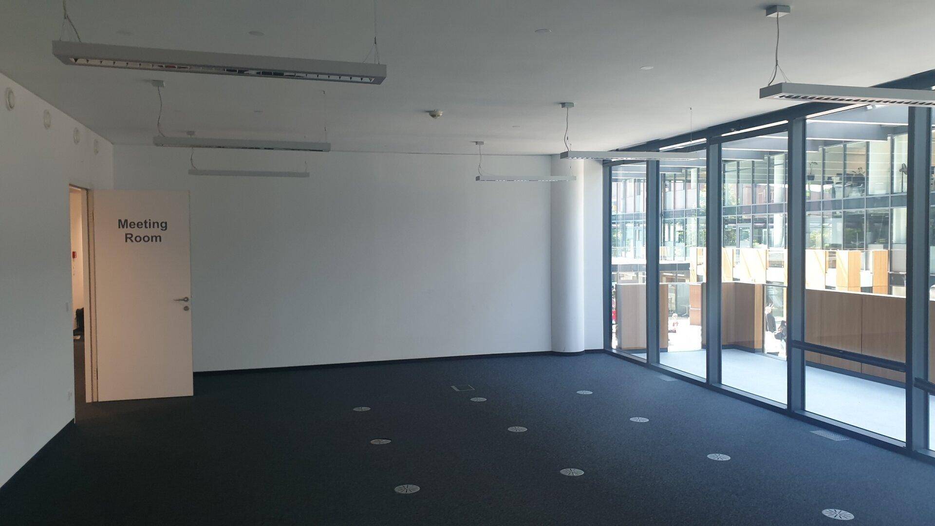 Leerer Bürobereich mit Zugang zu einem Besprechungsraum und großen Fensterfronten für viel Tageslicht.