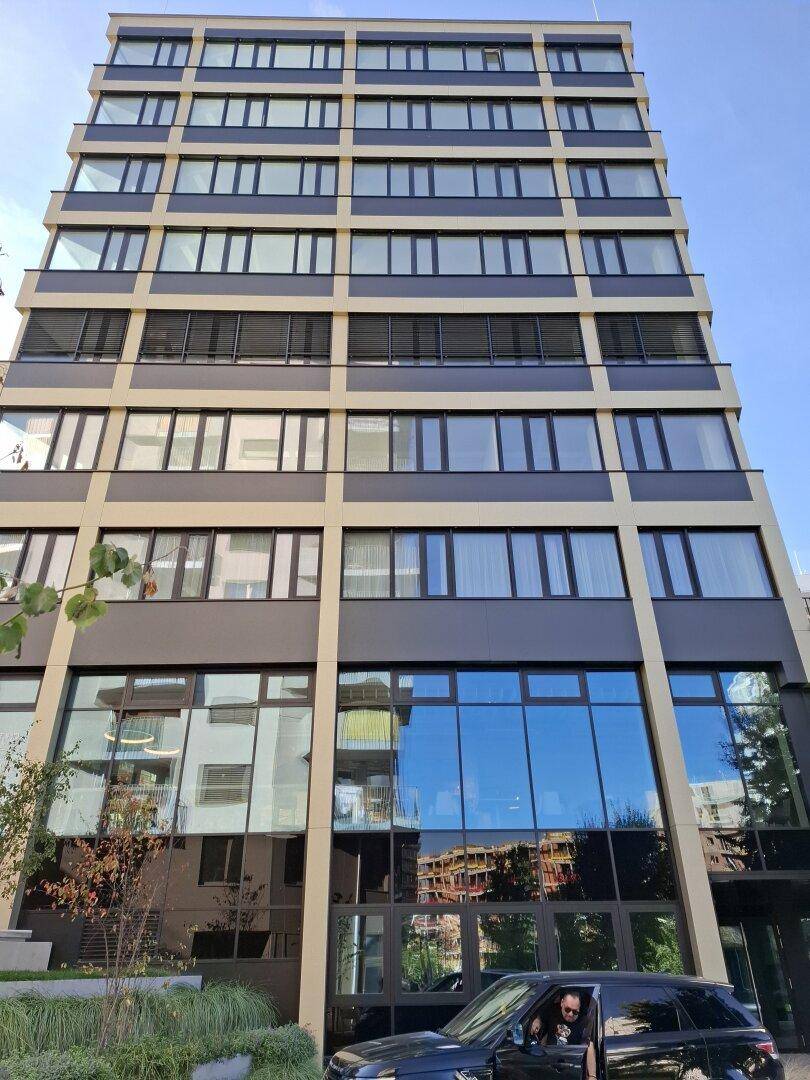 Hohes, modernes Gebäude mit reflektierender Glasfassade und einem geparkten Auto davor.