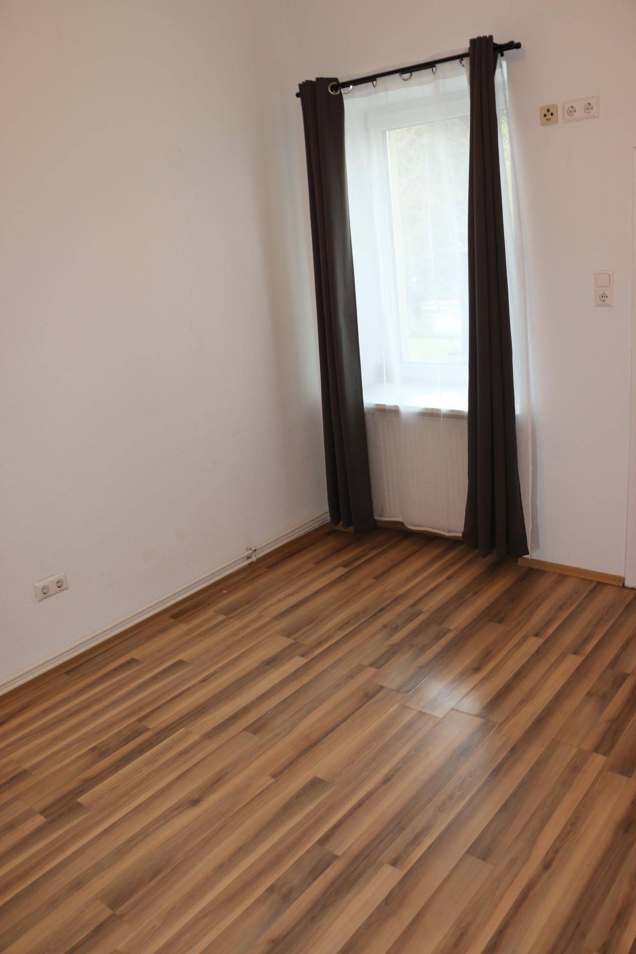 Helles Zimmer mit Laminatboden, Fenster und dunklen Vorhängen.