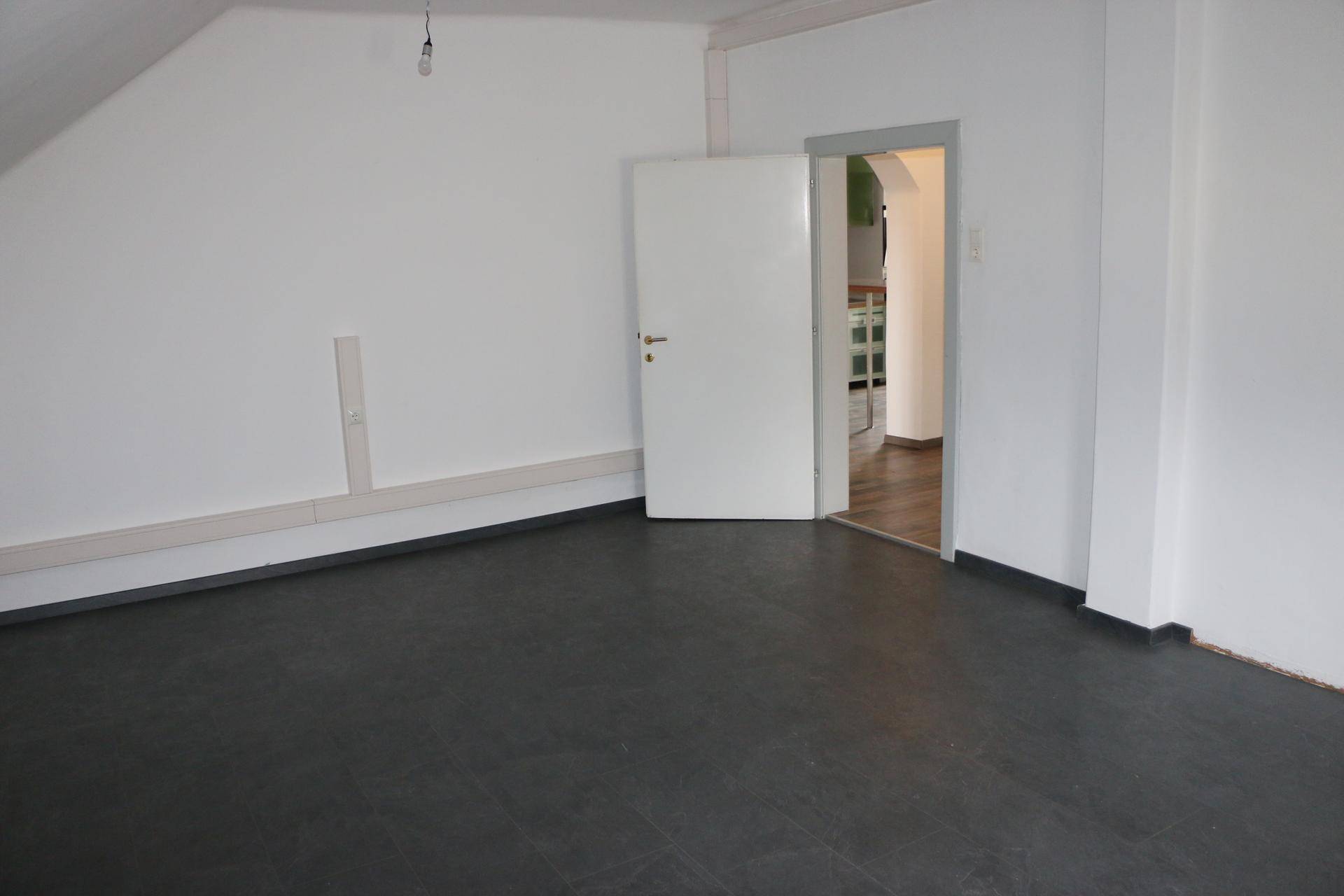 Unmöbliertes Dachzimmer mit dunklem Boden und Zugang zu einem weiteren Raum.