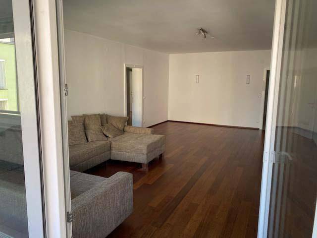 Geräumiges Wohnzimmer mit dunklem Holzboden und Blick durch die Balkontür nach draußen.