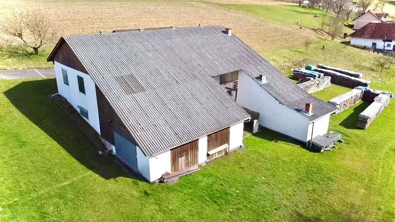 Luftaufnahme eines großen Nebengebäudes mit Wellblechdach, umgeben von Grünflächen und Holzstapeln.