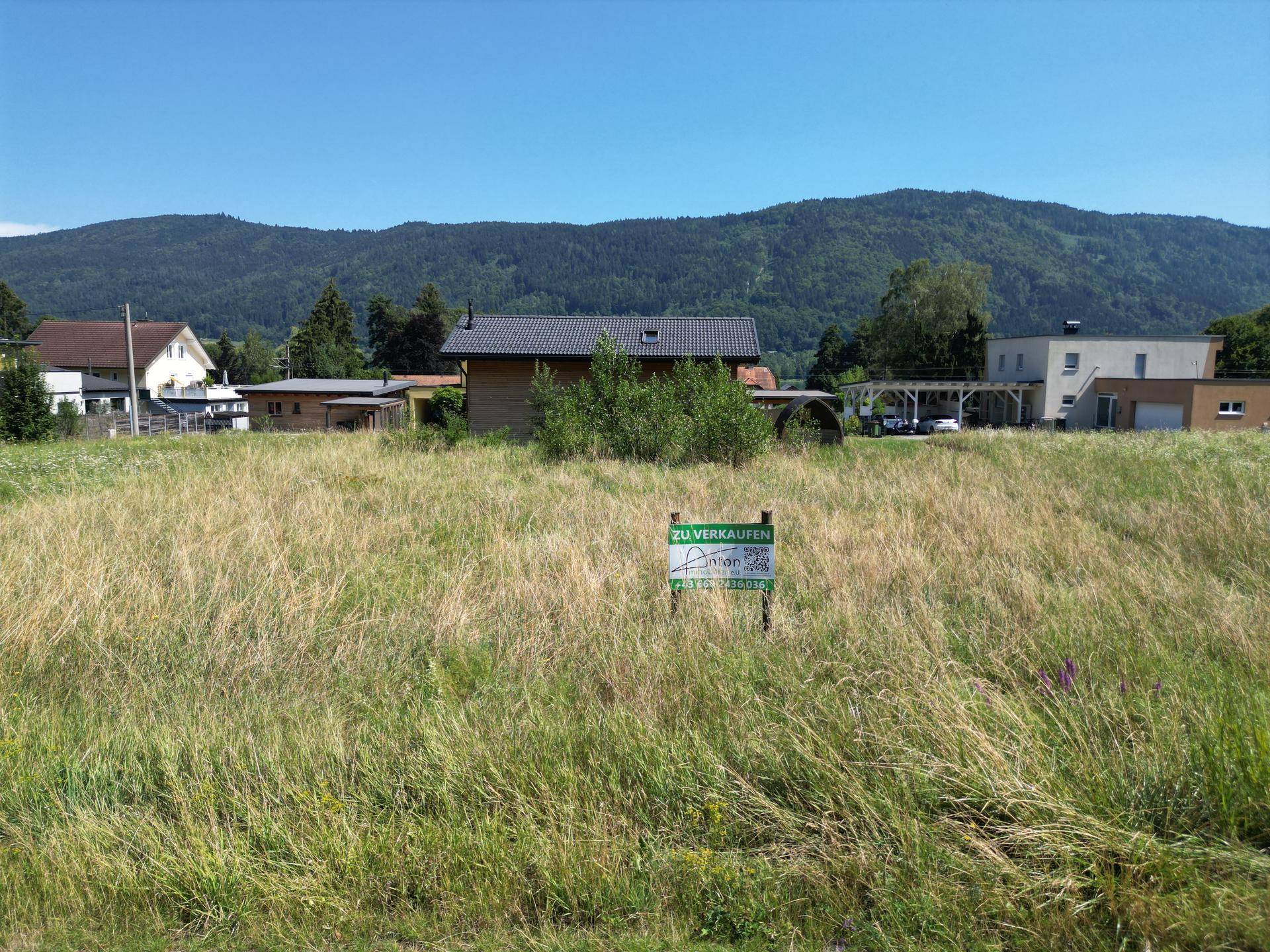 Grundstück mit 'Zu verkaufen'-Schild, umgeben von Natur und Bergen im Hintergrund.